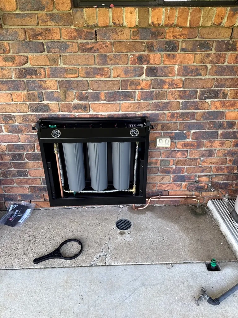Whole-house triple water filter system installed in a black cabinet on a brick wall